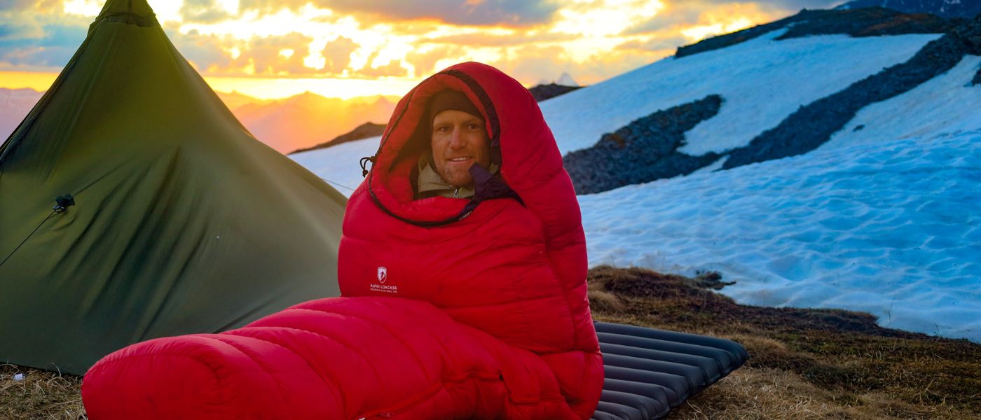 Ein Mann in einem Schlafsack neben einem Zelt auf einem verschneiten Berg, passend für Outdoor-Aktivitäten und Produkte von Alpinloacker.