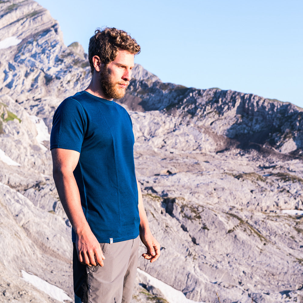 Ein Mann mit Bart in blauem Hemd steht vor einer Bergkulisse, passend zum Outdoor-Sortiment von alpinloacker.com.