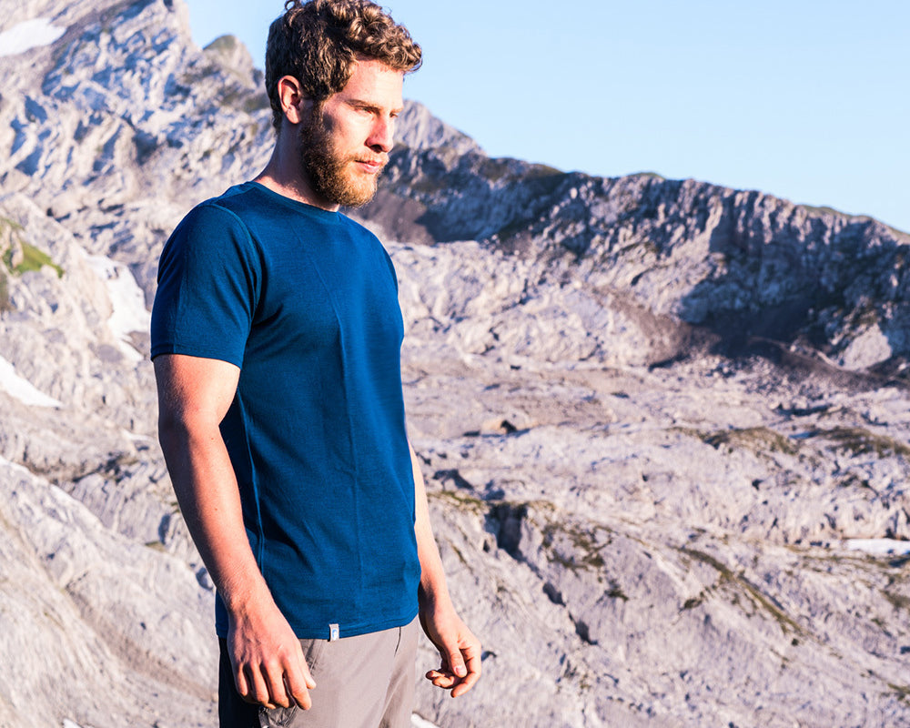 Ein Mann mit Bart in blauem Hemd steht vor einer Bergkulisse, passend zum Outdoor-Sortiment von alpinloacker.com.