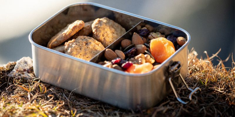 Eine Metallbox mit verschiedenen Lebensmitteln, darunter Obst, passend für Outdoor-Aktivitäten, angeboten von einem Outdoor-Shop spezialisiert auf Wanderausrüstung.