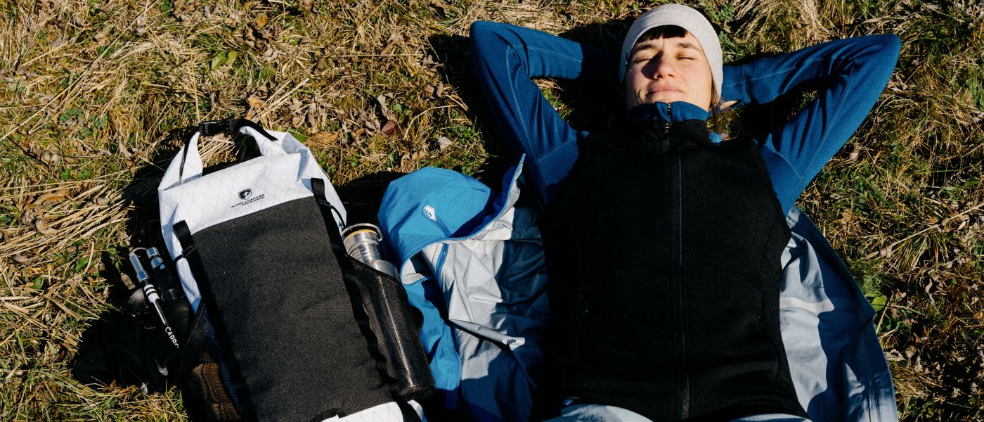 Eine Frau liegt im Gras mit einem schwarz-weißen Rucksack neben sich, passend für Outdoor-Aktivitäten wie Wandern.