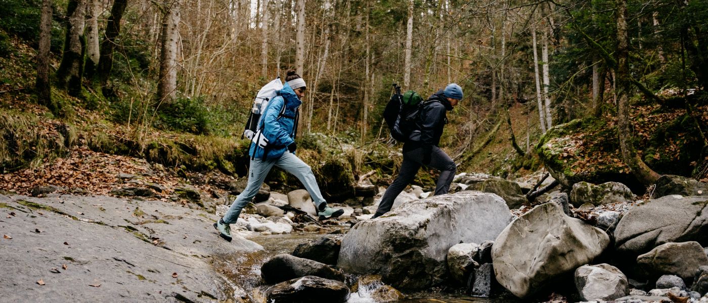 Ein Mann und eine Frau wandern über Felsen in einem Wald, beide tragen Outdoor-Bekleidung und Rucksäcke, passend für Alpinloacker-Ausrüstung.