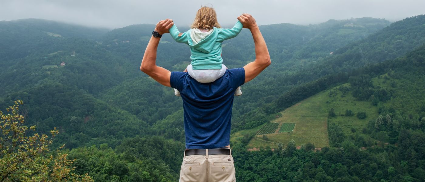 Ein Mann trägt ein Kind auf den Schultern beim Wandern, umgeben von Natur, passend zu Outdoor-Ausrüstung von alpinloacker.com.