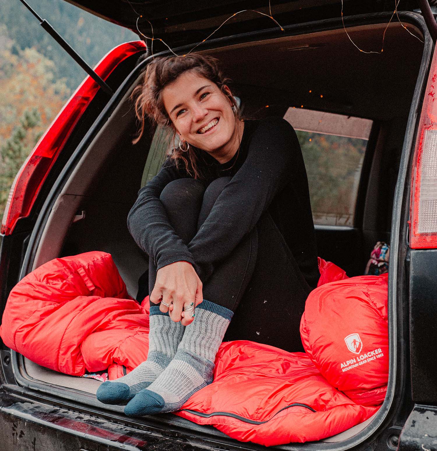 Eine Frau sitzt lächelnd im Auto, trägt graue Socken und hält eine rote Tasche mit Logo.