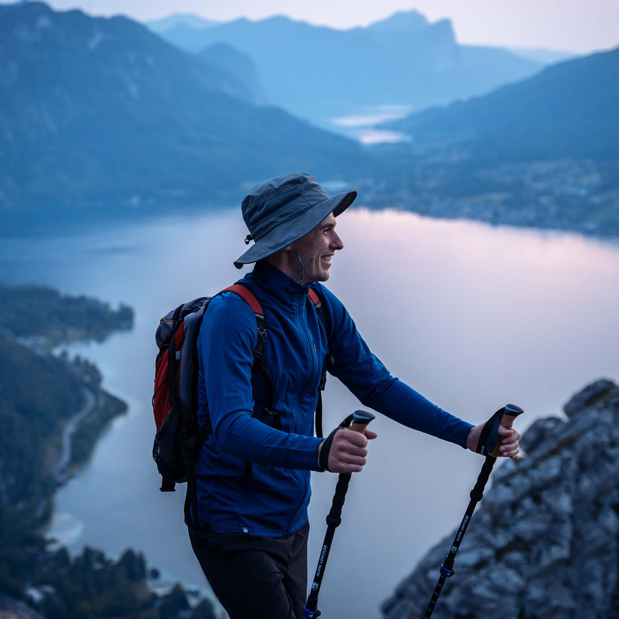 Ein Mann mit Hut und Rucksack wandert mit Stöcken auf einem Berg, im Hintergrund ein See und Berge.