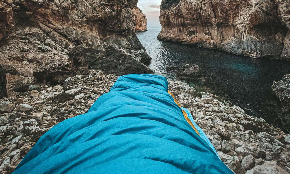 Ein blauer Schlafsack liegt auf einem felsigen Ufer mit Blick auf ein Gewässer, passend für Outdoor-Aktivitäten und Camping.
