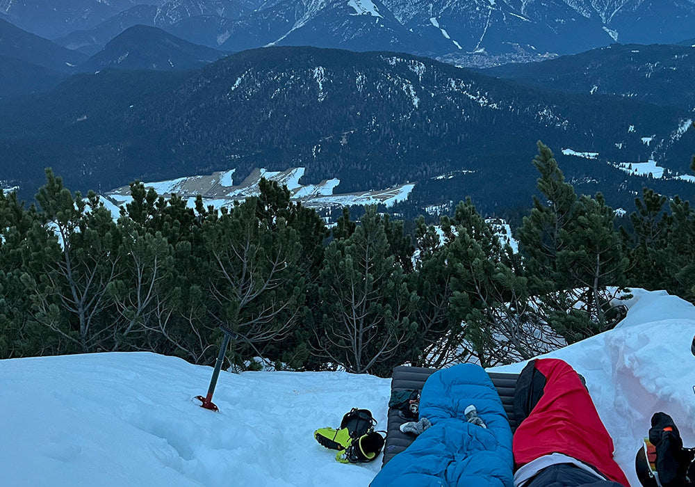 Winterbiwak in den Bergen bei Dämmerung: Zwei Schlafsäcke (blau, rot) auf Isomatten, umgeben von Bergstiefeln, Helmen und einem Eispickel im Schnee.