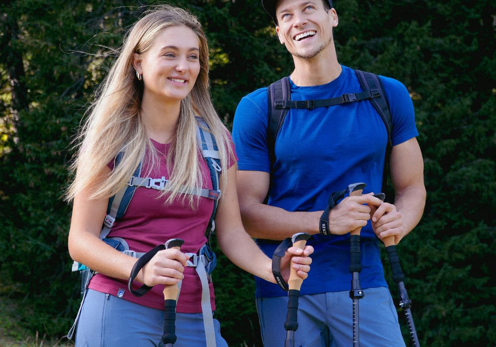 Zwei Personen beim Wandern mit Trekkingstöcken und leichten Outdoor-Kleidern in einer Waldlandschaft, repräsentierend Alpinloacker's Sortiment an Outdoor-Ausrüstung.