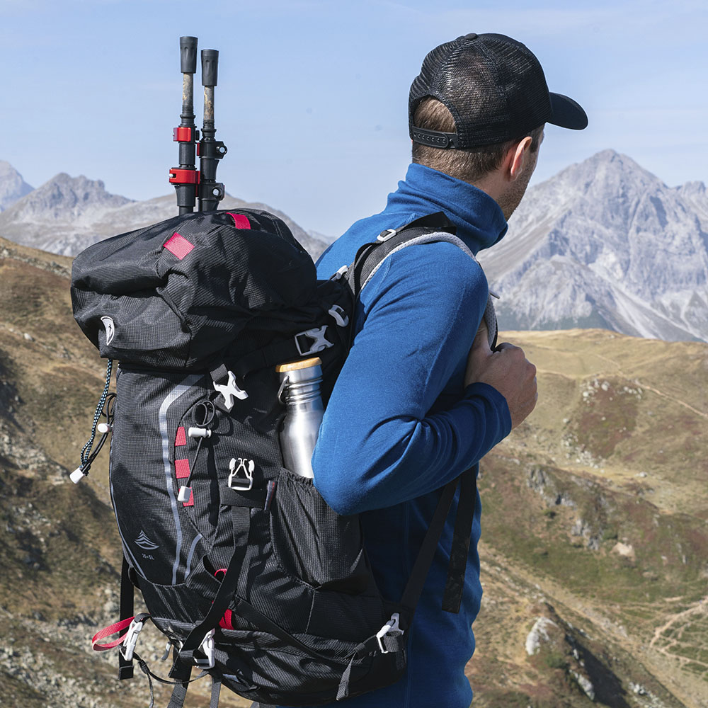 Hiker mit Alpin Loacker 35-Liter Wanderrucksack, Trekkingstöcke und Metallflasche sichtbar, blickt auf bergige Landschaft. Ideal für Outdoor-Abenteuer, robust und umweltfreundlich.