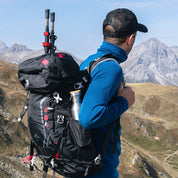 Hiker mit Alpin Loacker 35-Liter Wanderrucksack, Trekkingstöcke und Metallflasche sichtbar, blickt auf bergige Landschaft. Ideal für Outdoor-Abenteuer, robust und umweltfreundlich.