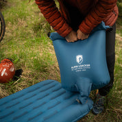 Person nutzt blauen Alpin Loacker Pump-Sack, um blaue Leichte Isomatte für 4 Jahreszeiten auf Gras aufzublasen, neben Fahrrad und rotem Beutel.