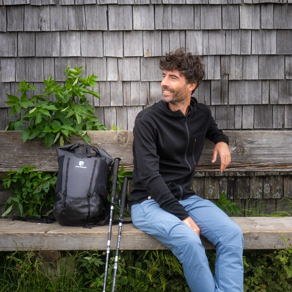 Mann auf Holzbank trägt 100% Merino Sport Hoody Zip Herren - 230 g/m2, mit schwarzem Rucksack und Wanderstöcken, passend für Outdoor-Abenteuer.
