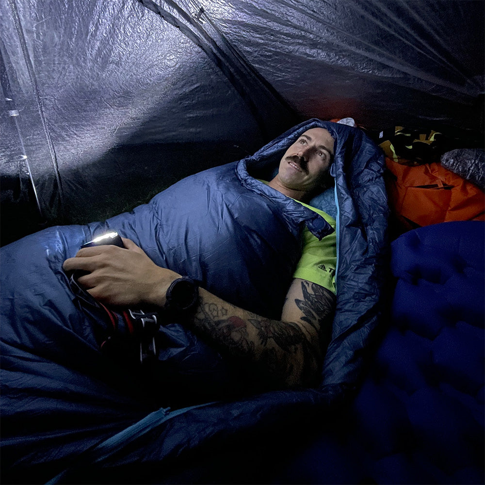 Mann im Zelt mit UL Down Pro 3 Daunenschlafsack, liegend auf Schlafmatte, hält Lichtquelle, umgeben von Campingausrüstung. Perfekt für Leichtigkeit und Nachhaltigkeit beim Campen.