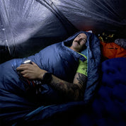 Mann im Zelt mit UL Down Pro 3 Daunenschlafsack, liegend auf Schlafmatte, hält Lichtquelle, umgeben von Campingausrüstung. Perfekt für Leichtigkeit und Nachhaltigkeit beim Campen.