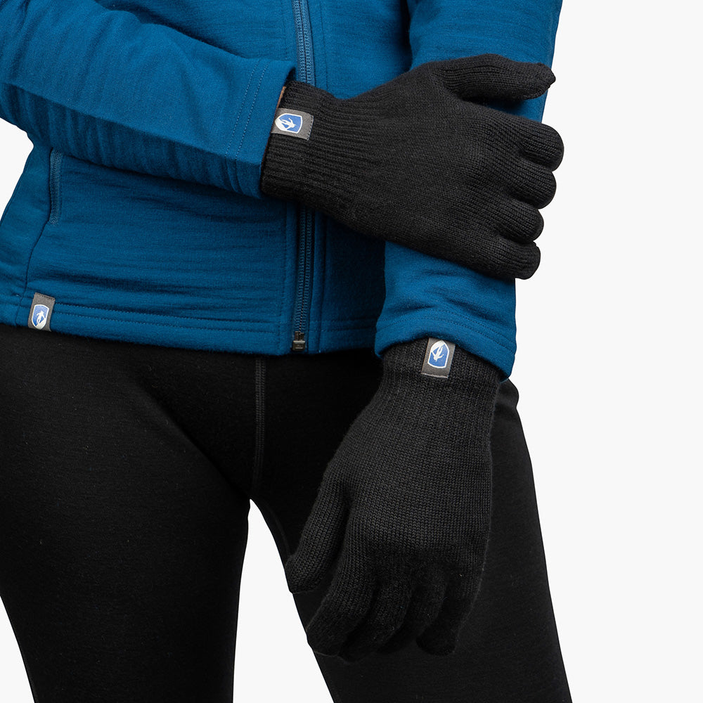 Person trägt blaue Jacke, schwarze Merino Handschuhe Damen & Herren mit Logo, Arme verschränkt. Perfekt für Outdoor-Abenteuer, in klassischer und fingerloser Ausführung erhältlich.
