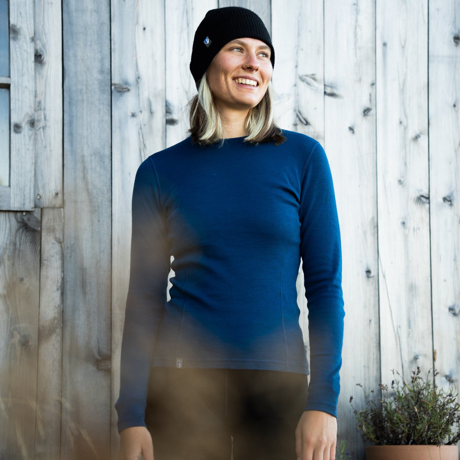 Frau steht in weichem 230g, blau-schwarzem Merino Set vor einer Holzwand