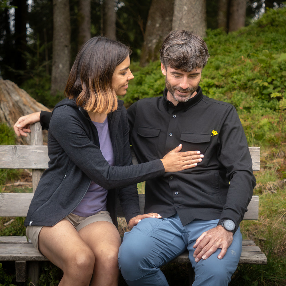 Wanderpärchen sitzt auf einer Bank in der Natur. Die Frau streicht über das schwarze Merino Wanderhemd ihres Partners