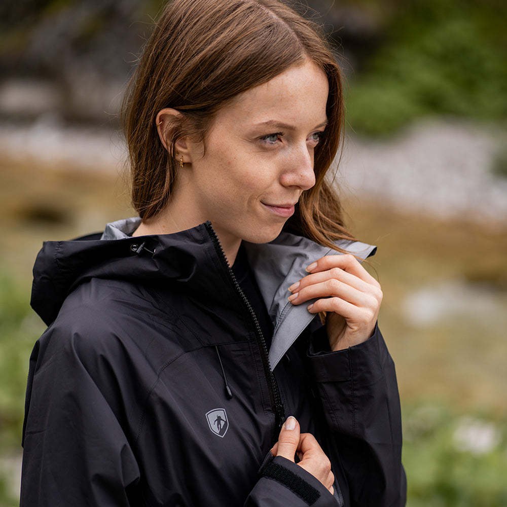 Junge Frau im Freien mit der 3 Lagen Damen Hardshell Regenjacke von Alpin Loacker, beim Anpassen des wasserdichten, atmungsaktiven schwarzen Hardshells mit Kapuze.