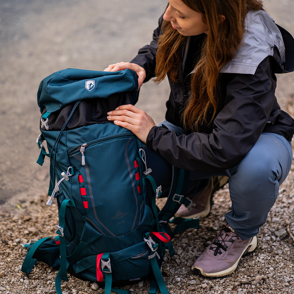 Person mit 35 Liter Wanderrucksack Herren & Damen von Alpin Loacker, verstellbarem Tragesystem, öffnet den oberen Bereich während einer Outdoor-Aktivität auf kiesigem Untergrund.