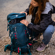 Person mit 35 Liter Wanderrucksack Herren & Damen von Alpin Loacker, verstellbarem Tragesystem, öffnet den oberen Bereich während einer Outdoor-Aktivität auf kiesigem Untergrund.