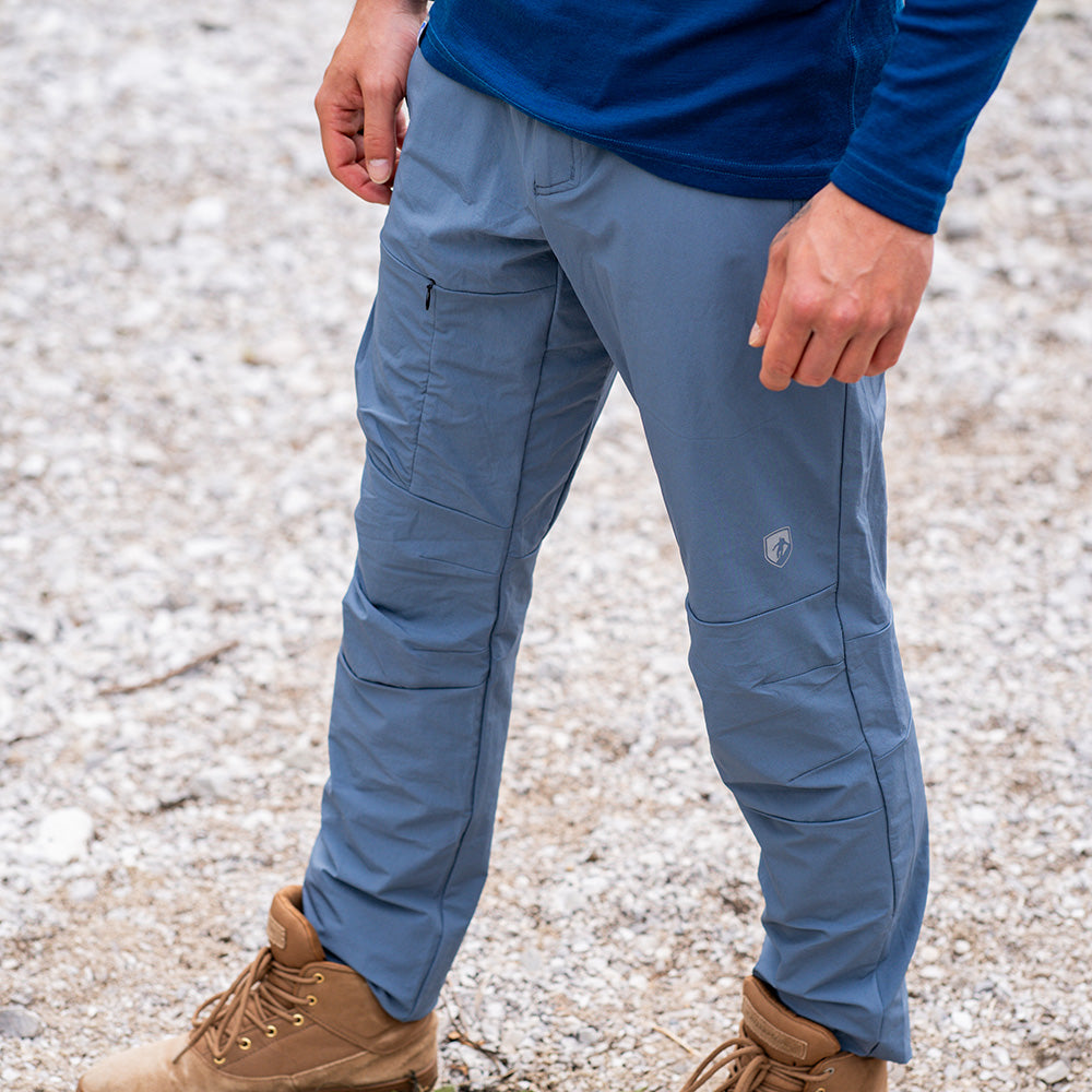 Softshell Wanderhose Herren auf steinigem Untergrund, zeigt Reißverschlusstasche am Oberschenkel, kombiniert mit beigen Wanderschuhen. Geeignet für Trekking mit Komfort und Flexibilität.
