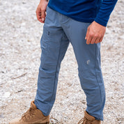 Softshell Wanderhose Herren auf steinigem Untergrund, zeigt Reißverschlusstasche am Oberschenkel, kombiniert mit beigen Wanderschuhen. Geeignet für Trekking mit Komfort und Flexibilität.
