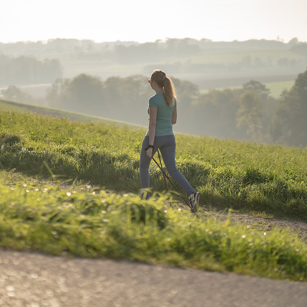 Frau mit Teleskop Nordic Walking Stöcke aus Aluminium von Alpin Loacker, wandert auf einem Pfad durch sanfte Hügel, umgeben von Baumlinien in der Ferne.