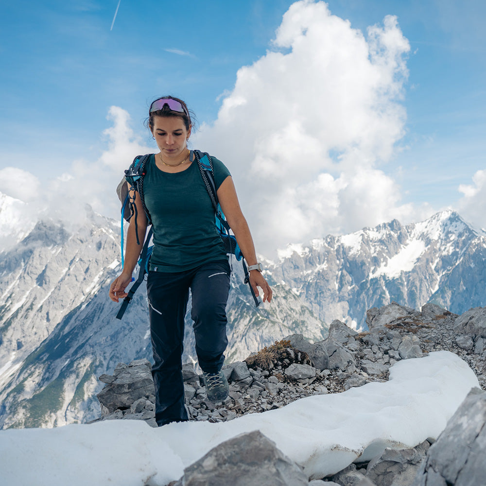 Hikerin mit Rucksack trägt das Alpin Loacker 100% Merino T-Shirt Damen 180g/m2 beim Aufstieg über schneebedeckten Bergkamm, umgeben von alpinen Gipfeln.
