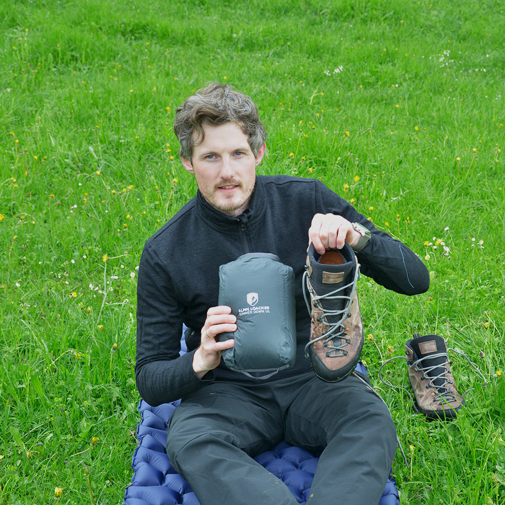 Mann zeigt Alpin Loacker Ultra leichter Daunen Sommerschlafsack 290g im Kompressionssack neben abgenutztem Wanderschuh, betont Leichtigkeit und Kompaktheit für Campingabenteuer.