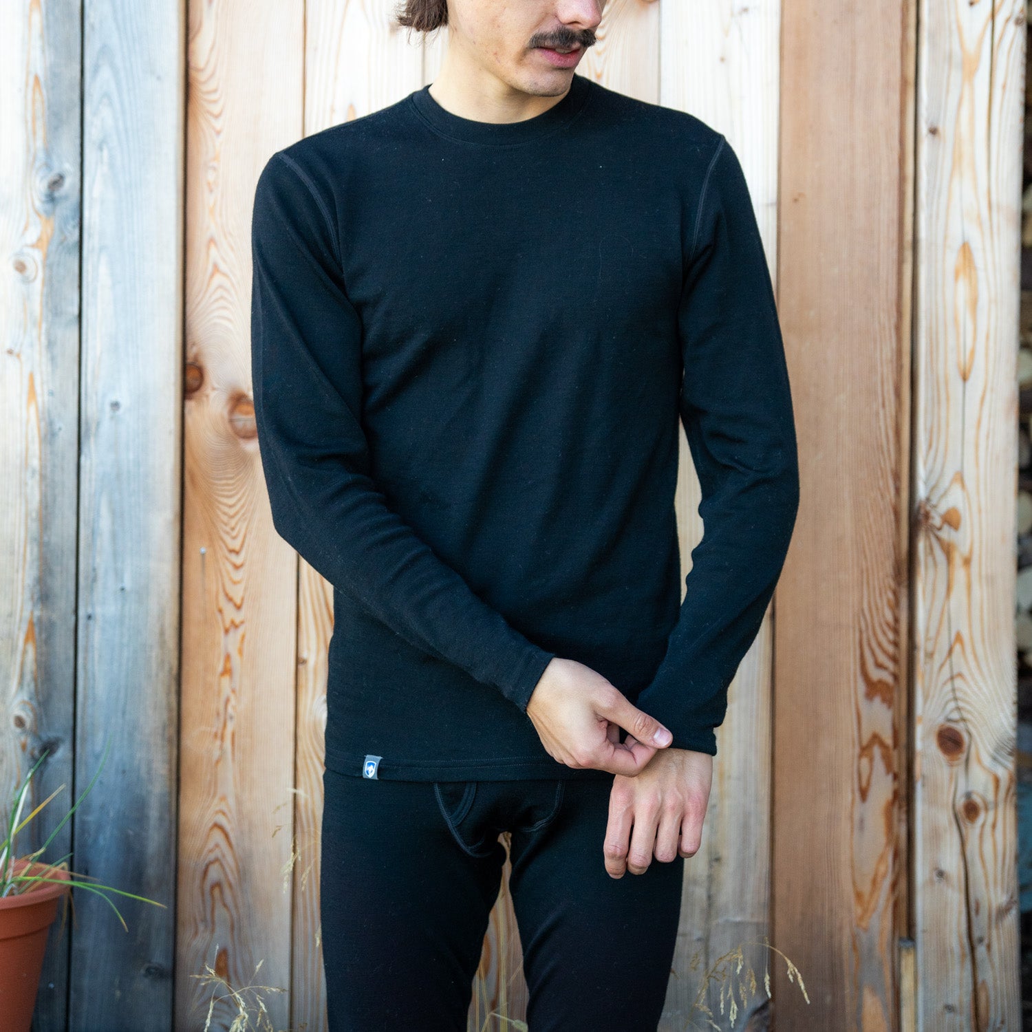 Mann trägt das 100% Merinowolle Set schwarz von Alpin Loacker vor einer Holzwand, perfekt als Baselayer für die kalte Jahreszeit