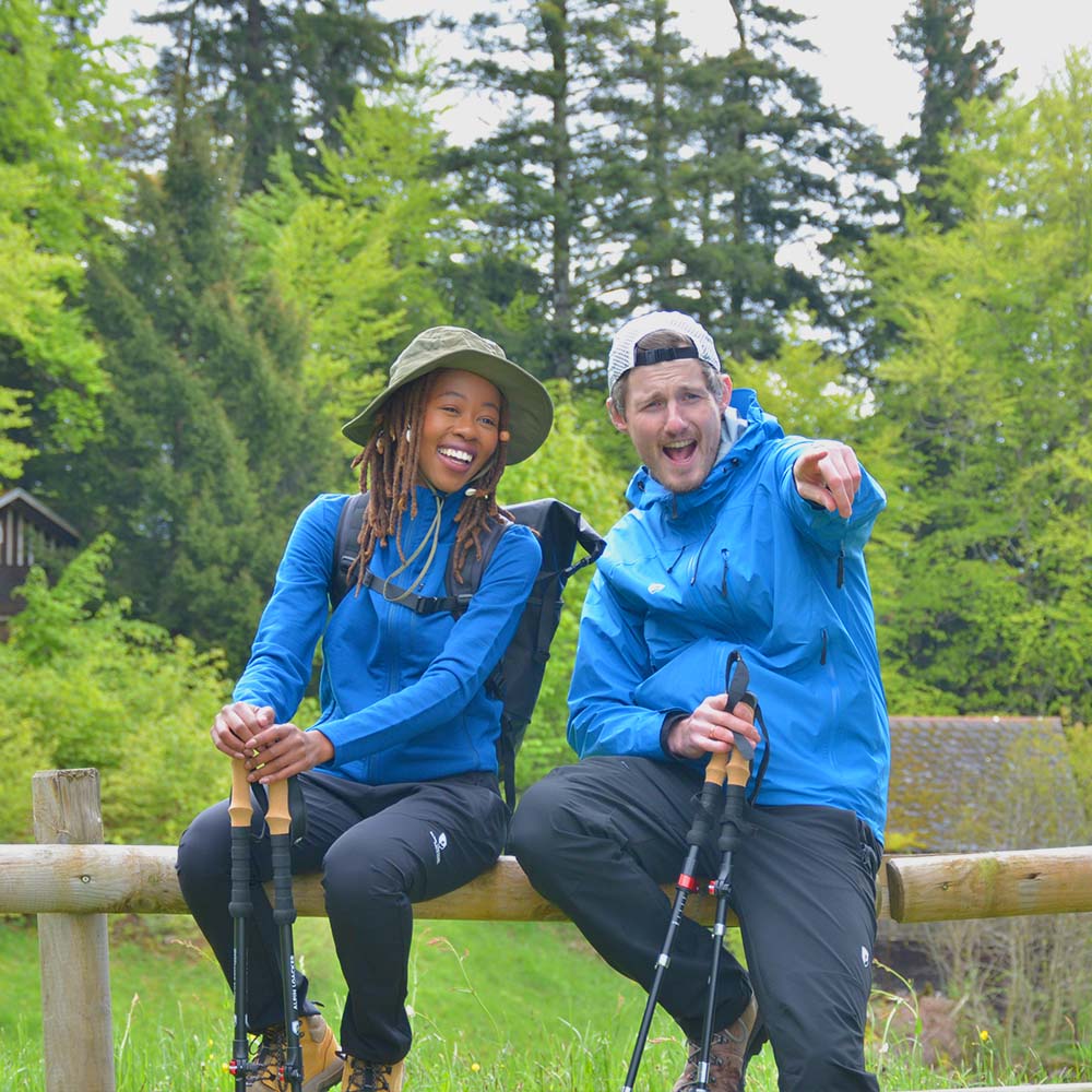 Zwei Wanderer im Wald, tragen Softshell Wanderhose Herren. Sie pausieren auf einem Zaun, halten Trekkingstöcke. Perfekt für Outdoor-Touren, leicht, wasserabweisend, flexibel.