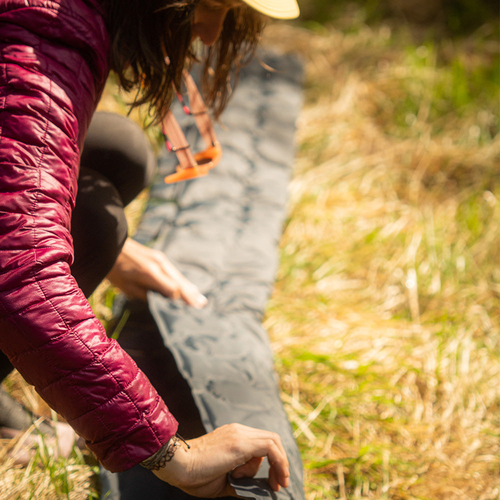 Person glättet graue Ultra leichte Isomatte Air Light im Freien, trägt maroonfarbene Jacke. Ideal für Camping, aus 100% recyceltem RipStop Nylon, kompakt und leicht.