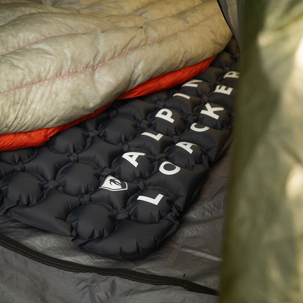 Nahaufnahme der Ultra leichten Isomatte Air Light im Zelt, mit einem Schlafsack darauf, zeigt das ALPINE PACKER-Logo und die wabenförmige Struktur für optimale Isolation.