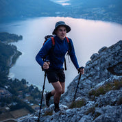 Wanderer mit Merino Jacke Herren 270 g/m² erklimmt steinigen Berg, trägt Trekkingstöcke und Rucksack, vor ruhigem Bergsee und bewaldeter Landschaft.