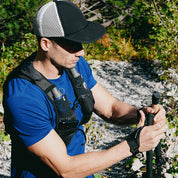 Mann justiert Faltbare Trailrunning Stöcke aus Carbon auf einem felsigen Waldweg, trägt Hydrationsweste und Baseballkappe, bereit für anspruchsvolle Outdoor-Aktivitäten.