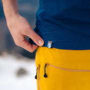 Nahaufnahme eines Merino Langarmshirt Herren 230 g/m2 in einem verschneiten Outdoor-Setting. Das Shirt wird von einer Hand gehalten, passende gelbe Outdoor-Hose erkennbar.