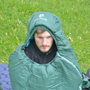 Person im SYN PRO - Synthetik Sommer Schlafsack ultraleicht von ALPIN LOACKER auf blauer Campingunterlage, umgeben von Wildblumen, mit kleinem Rucksack daneben.
