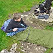 Mann im Bivi PRO - leichter Biwaksack, im Freien auf einem Schlafpad, umgeben von Wanderausrüstung wie Stöcken, Rucksack und Stiefel. Ideal für Outdoor-Abenteuer.