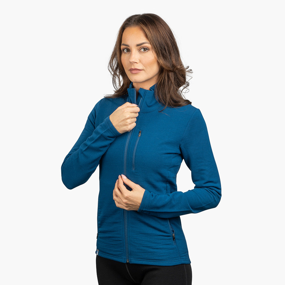 Frau trägt Merino Jacke Damen 270 g/m2, passt den Reißverschluss an. Die Jacke betont Sportlichkeit und Funktionalität, ideal für Outdoor-Abenteuer.