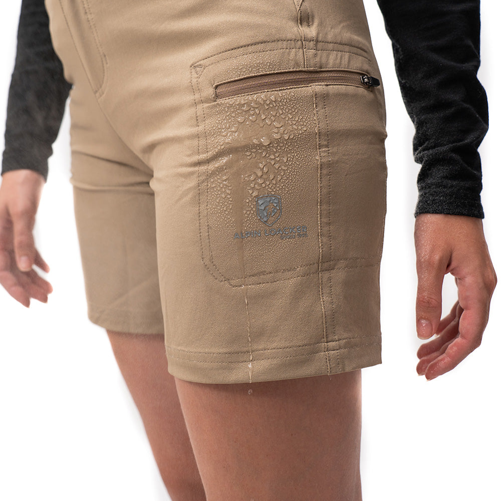 Kurze Wanderhose Damen: Nahaufnahme einer wasserabweisenden, hell-khakifarbenen Shorts mit Reißverschlusstasche und sichtbar abperlendem Wasser, getragen von einer Person.