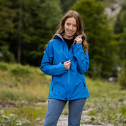 Frau in blauer 3 Lagen Damen Hardshell Regenjacke auf Pfad, lächelnd, hält Reißverschluss und Kragen. Ideal für Outdoor-Abenteuer, wasserdicht und atmungsaktiv.