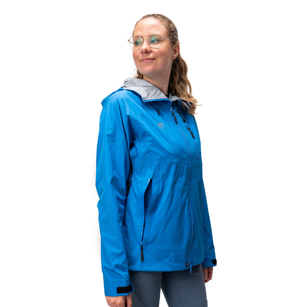 Person trägt eine 3 Lagen Damen Hardshell Regenjacke in Blau mit Kapuze, Reißverschlusstaschen und Klettverschlüssen an den Handgelenken, ideal für Outdoor-Aktivitäten.