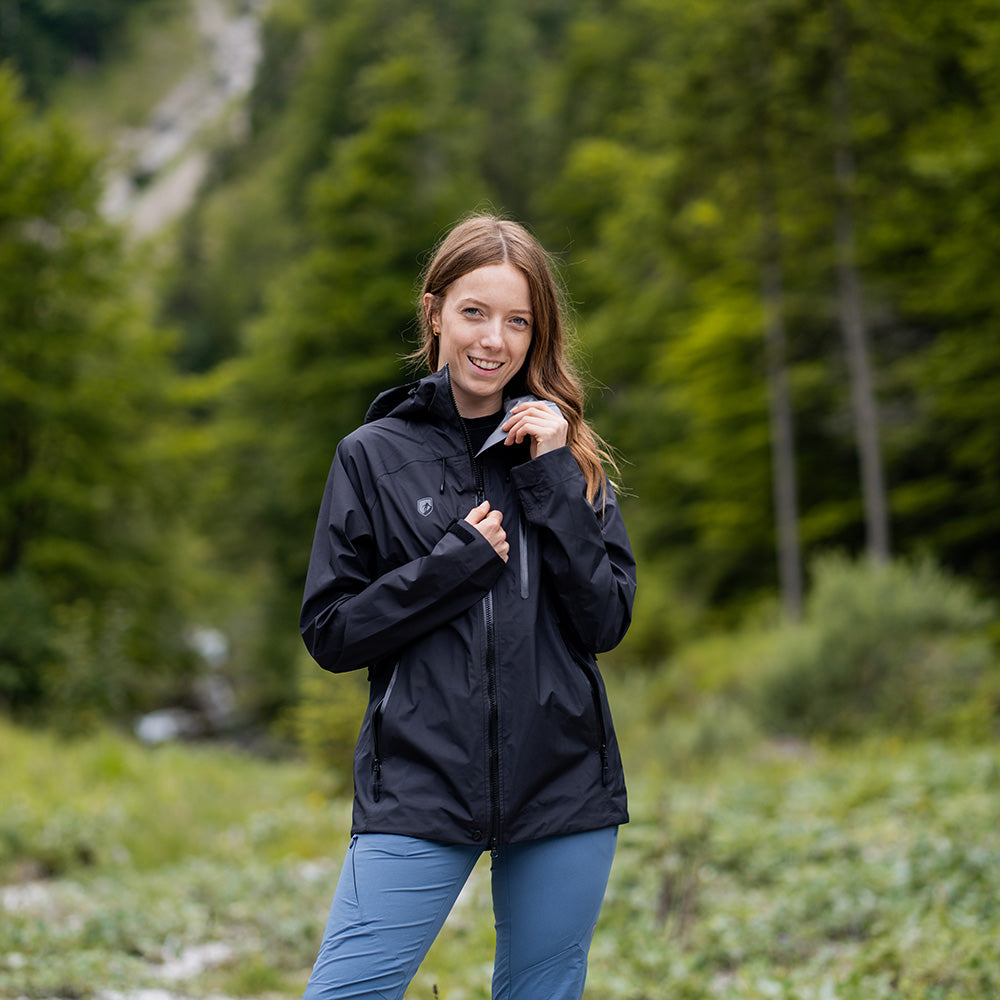 Junge Frau in Wald mit 3 Lagen Damen Hardshell Regenjacke, ideal für Outdoor-Aktivitäten, trägt Helm-kompatible Kapuze und verstellbaren Kragen, zeigt Vielseitigkeit und Schutz.