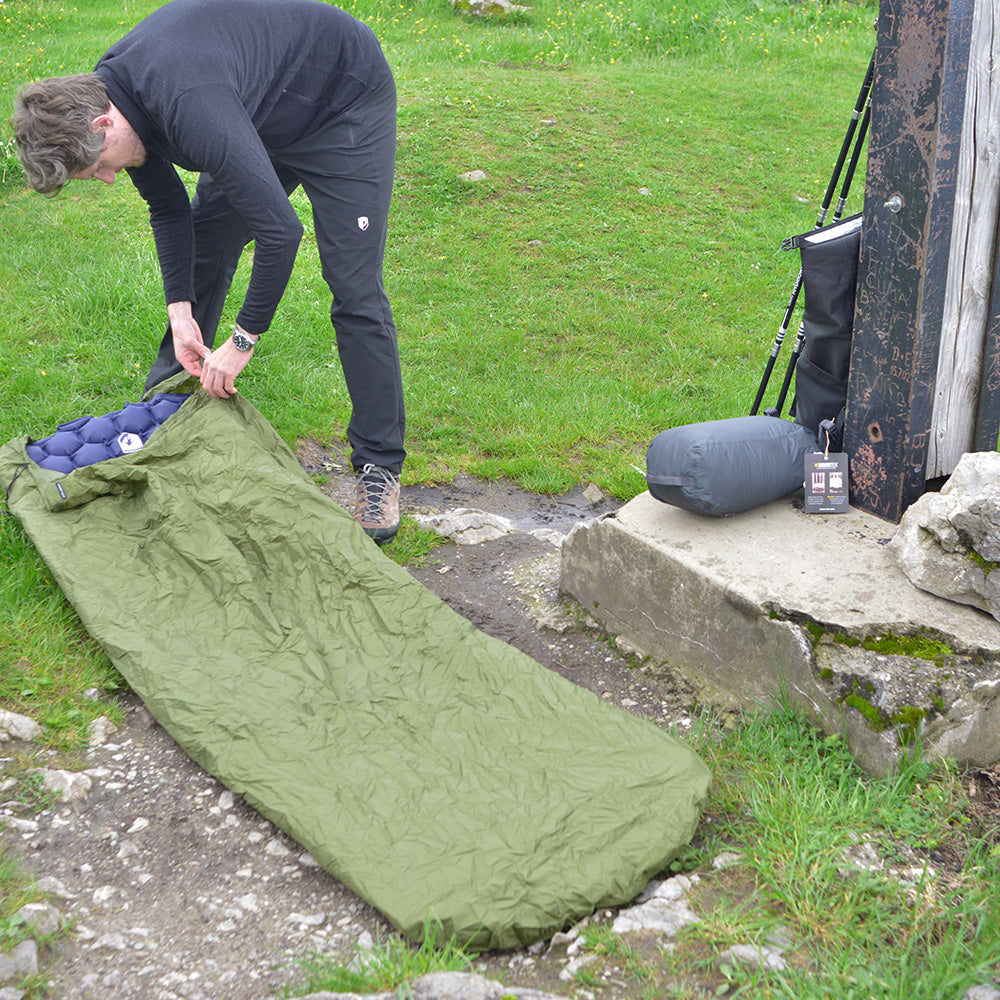 Camper richtet Bivi PRO - leichter Biwaksack aus RipStop Nylon auf Gras ein, Trekkingstöcke und gepacktes Zelt im Hintergrund, für Outdoor-Abenteuer.