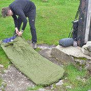 Camper richtet Bivi PRO - leichter Biwaksack aus RipStop Nylon auf Gras ein, Trekkingstöcke und gepacktes Zelt im Hintergrund, für Outdoor-Abenteuer.