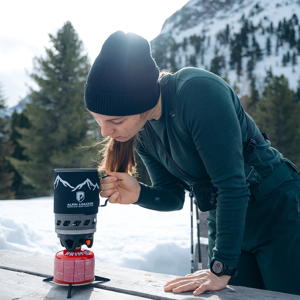 Person kocht mit dem Alpin Loacker Camping Gaskocher Set auf einem Picknicktisch, umgeben von Schnee und Bergen. Perfekt für Outdoor-Abenteuer und effizientes Kochen.