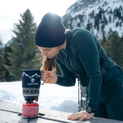 Person kocht mit dem Alpin Loacker Camping Gaskocher Set auf einem Picknicktisch, umgeben von Schnee und Bergen. Perfekt für Outdoor-Abenteuer und effizientes Kochen.