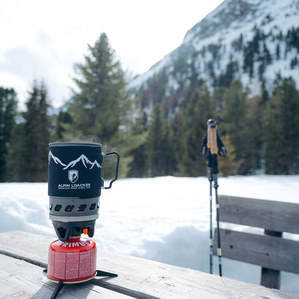 Camping Gaskocher Set auf einem Holztisch im Schnee, mit dampfendem Topf. Trekkingstöcke im Hintergrund, ideal für Outdoor-Abenteuer im Gebirge.