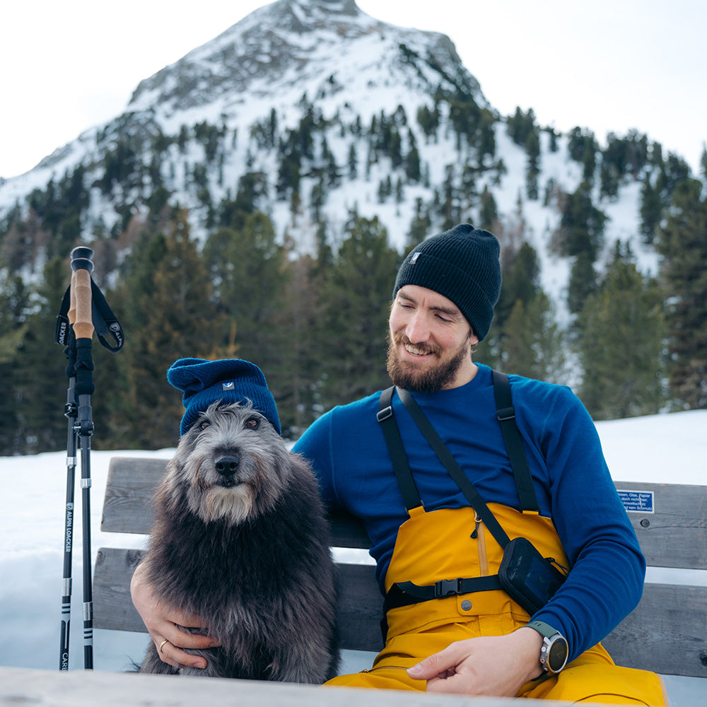 m² sitzt mit Hund und Stöcken vor schneebedecktem Berg, betont Wärme und Vielseitigkeit.