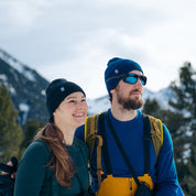 Zwei Personen in winterlicher Berglandschaft tragen die Merino Mütze Herren & Damen Strick 230g/m2, ideal für Outdoor-Abenteuer dank temperaturregulierender Merinowolle.